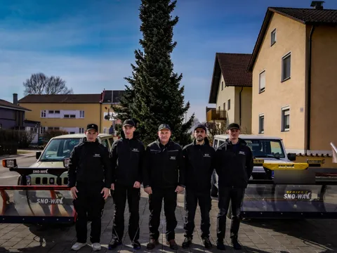 Fünfköpfiges MDZ-Team steht nebeneinander vor Mehrfamilienhäusern, im Hintergrund zwei mit Schneeschildern ausgerüstete Fahrzeuge für den Winterdienst.