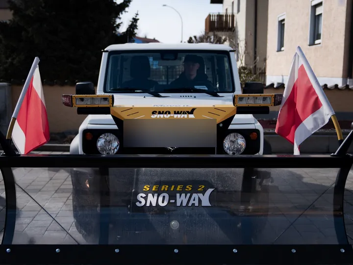 Frontansicht eines Winterdienst-Fahrzeugs mit Schneeschild und rot-weißen Warnfähnchen vor einer Wohnstraße in Regensburg.