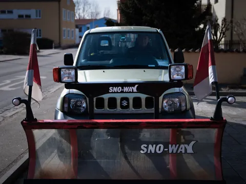 Winterdienst-Fahrzeug mit gelbem Schneeschild und Warnfähnchen, bereit für den Einsatz vor einem Wohngebäude.