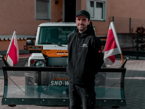 Lächelnder Mitarbeiter im MDZ-Jacket neben einem Winterdienst-Fahrzeug mit Schneeschild.