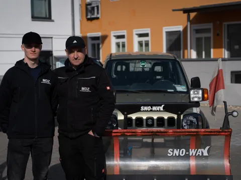Zwei Mitarbeiter unterschiedlichen Alters stehen gemeinsam vor einem Winterdienst-Fahrzeug mit Schneeschild.