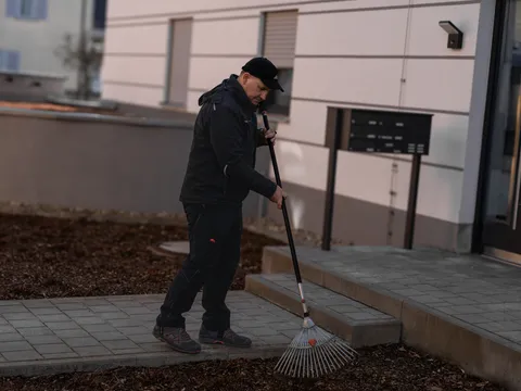 Mitarbeiter recht Laub auf dem Weg zum Hauseingang einer Wohnanlage zusammen.