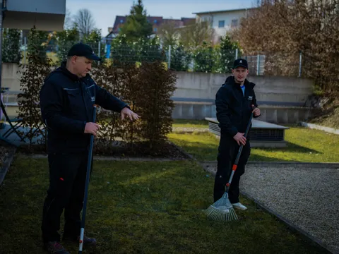 Zwei Mitarbeiter rechen gemeinsam den Rasen im Innenhof einer Wohnanlage.