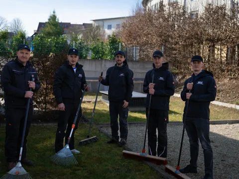 Fünfköpfiges MDZ-Team posiert mit Rechen und Besen im gepflegten Innenhofgarten einer Wohnanlage.