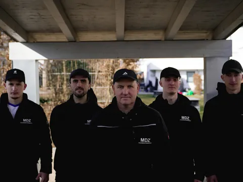 Fünfköpfiges MDZ-Team steht unter einem Pergola-Dach vor einer Wohnanlage.