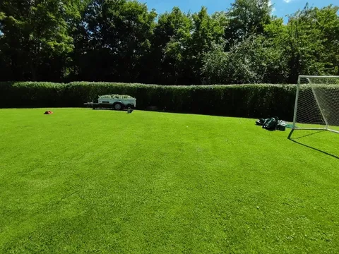 Große frisch gemähte Rasenfläche mit Fußballtor, dahinter eine sauber geschnittene Hecke und ein Anhänger voller Grünschnitt.